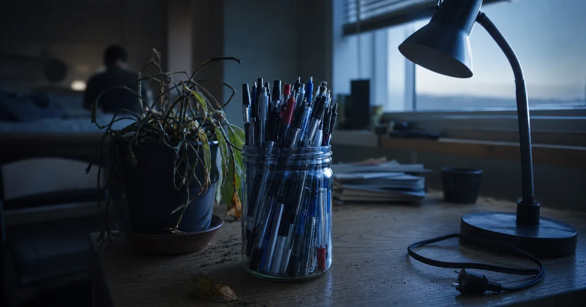 A cluttered desk at dawn in a shared student house, dozens of spent pens crowded into a jar beside a dying houseplant and an unplugged desk lamp, cold blue