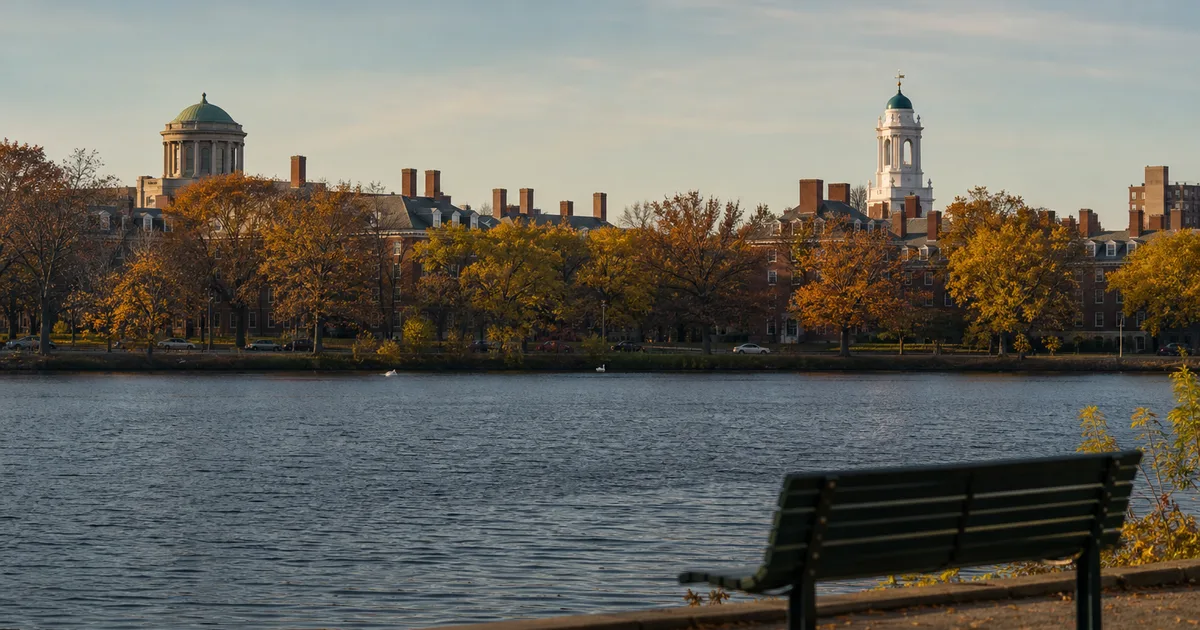 Autumn afternoon on the Charles River Esplanade with MIT and Harvard campus brick buildings visible across the water, golden foliage lining the riverbank