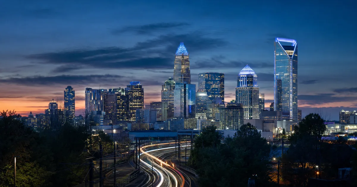 Glass-facade office towers lining Tryon Street in uptown Charlotte at dusk with warm interior lights visible through the windows