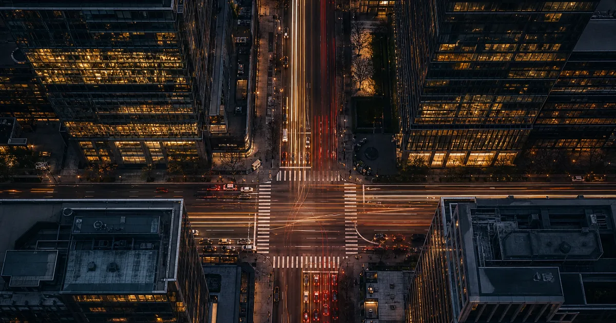 An aerial view of a busy crossroads in a downtown financial district at dusk, with illuminated glass office buildings on all four corners and light trails