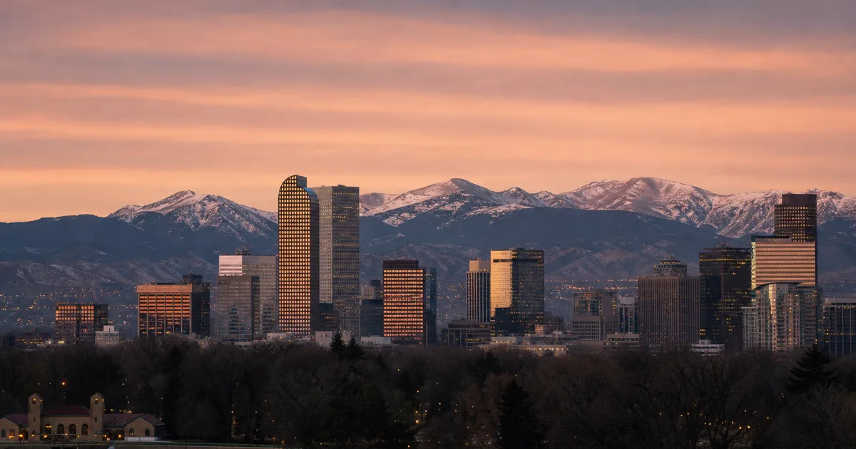 Downtown Denver skyline photographed at dusk from City Park, snow-capped Rocky Mountains as a dramatic backdrop behind glowing office towers