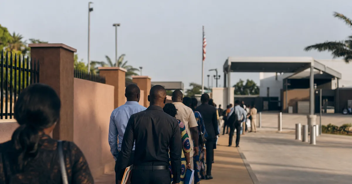 Exterior of a US diplomatic mission compound in a West African city, applicants with document folders waiting in a structured queue under morning sunlight