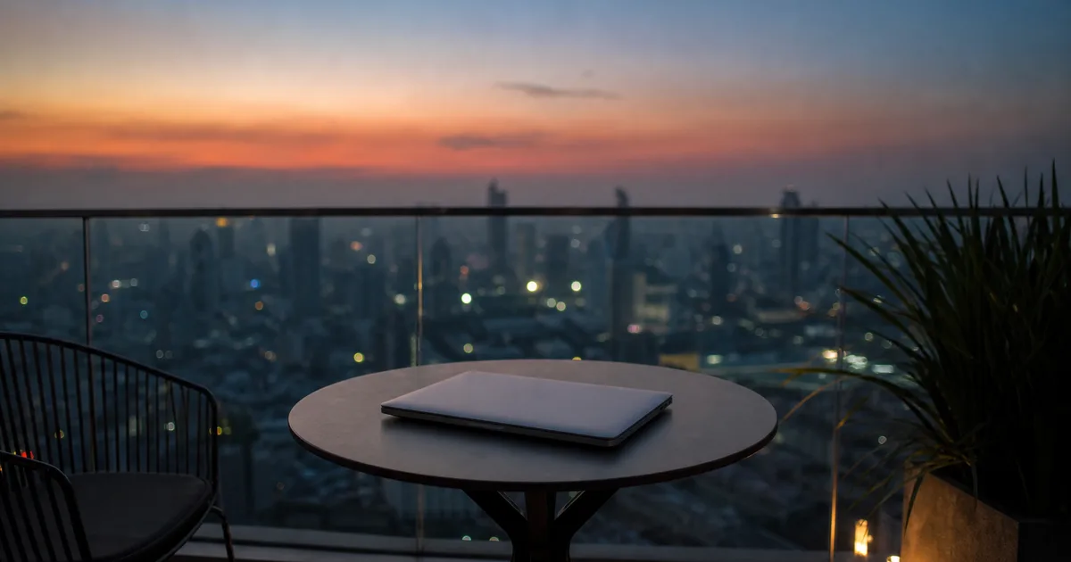 A rooftop apartment balcony at dusk, a small table with a closed laptop and a city skyline softening into evening haze