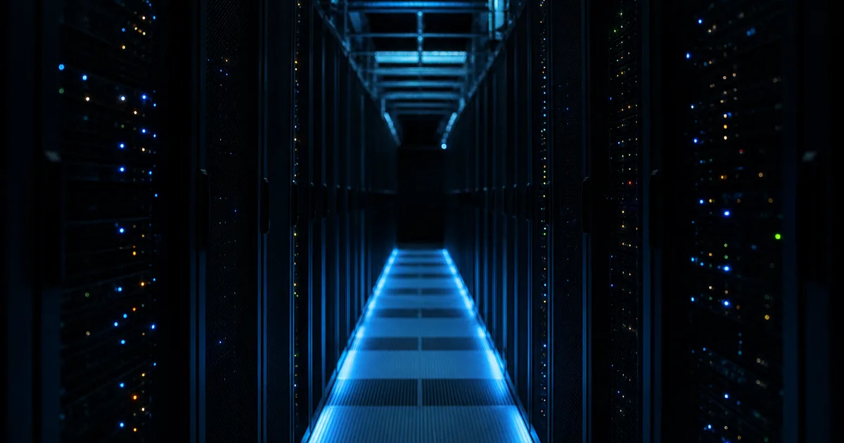 A dim server-room aisle at night with tall racks of humming equipment and rows of small indicator lights, cold blue glow washing a narrow metal walkway that