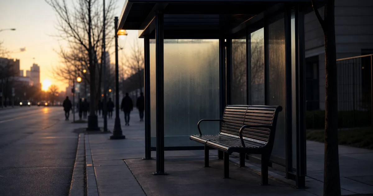 A city bus stop at dawn, an empty bench beneath a covered shelter with commuters' silhouettes distant down the block