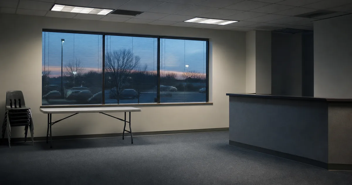 An empty suburban office suite at dusk with a folding table, stacked plastic chairs and a bare reception counter, cold overhead fluorescent light and