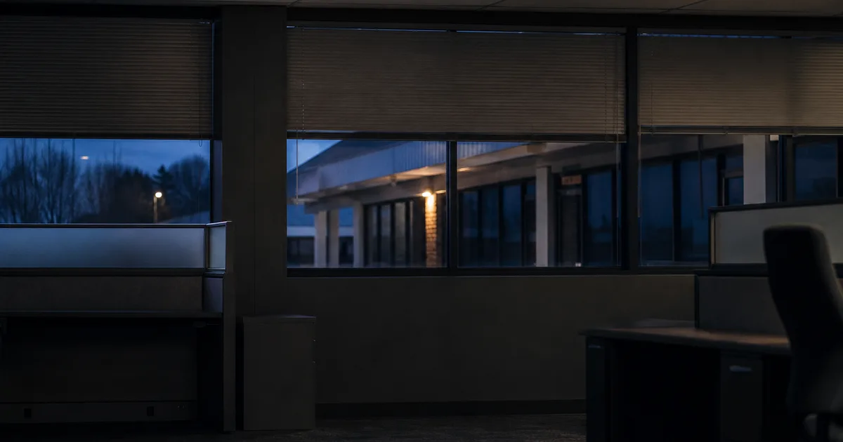 A student sits at a laptop late at night in a small apartment, papers scattered on the desk, city lights through the window.